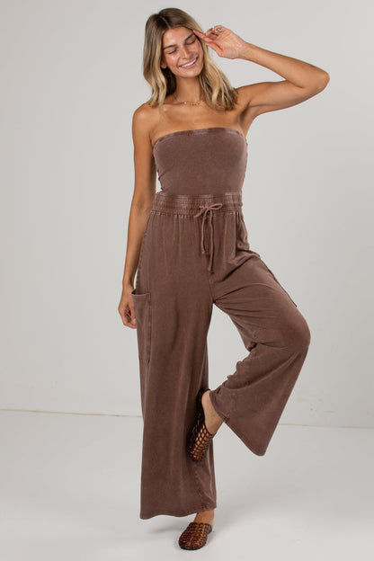 A woman modeling a brown strapless jumpsuit with wide-leg pants and a drawstring waist, photographed against a plain light gray studio background.