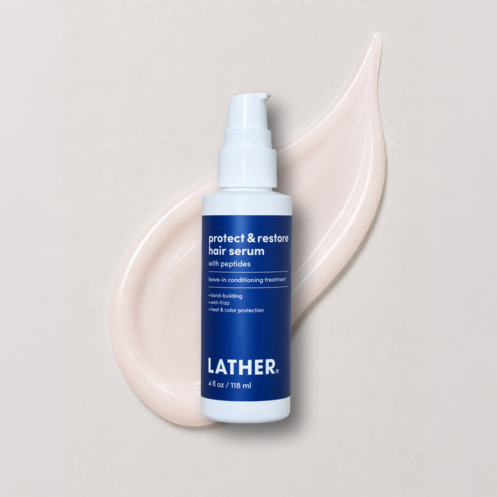 A bottle of Lather Protect & Restore Hair Serum, featuring a white pump bottle with a blue label, set against a beige background with product swatched behind it.