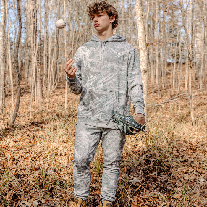 Person in light camouflage clothing holding a baseball and glove in a forest setting