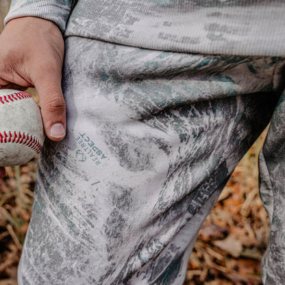 Person holding a baseball with camouflage-patterned pants in an outdoor setting