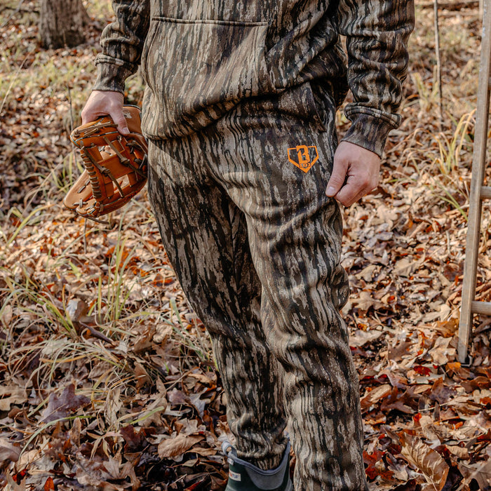 Person wearing camouflage hunting gear with a orange BL101 logo on pants standing in a forest.