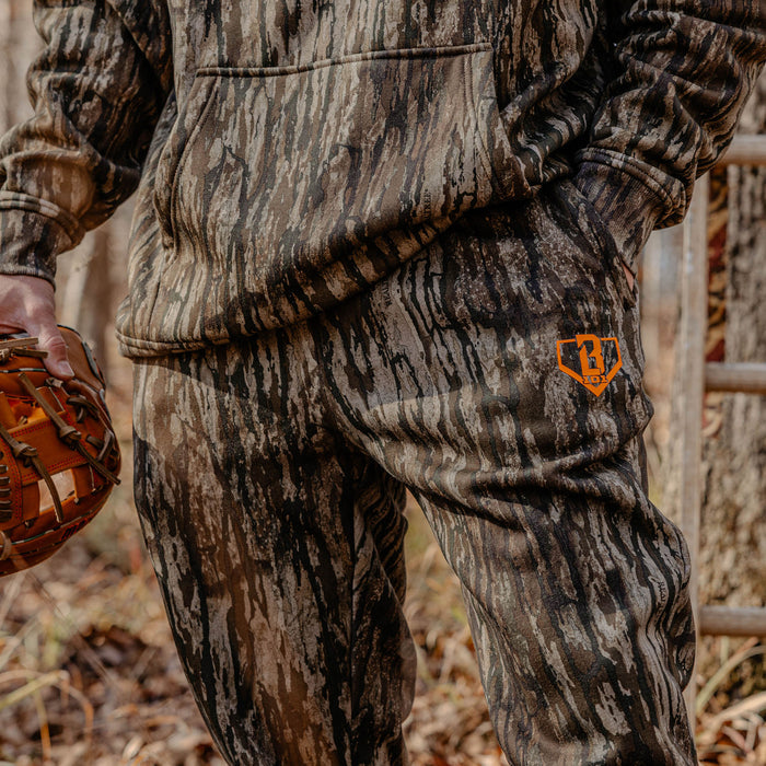 Person wearing camouflage hunting gear with a orange BL101 logo on pants, holding a baseball glove.