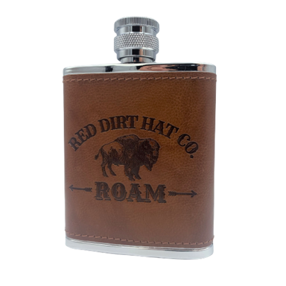 A bottle of Red Dirt Hat Company Roam cologne, with a brown leather-like wrap around the lower half and a metal cap. The bottle features the logo and the word 'ROAM' engraved on the front. It looks similar to a flask.