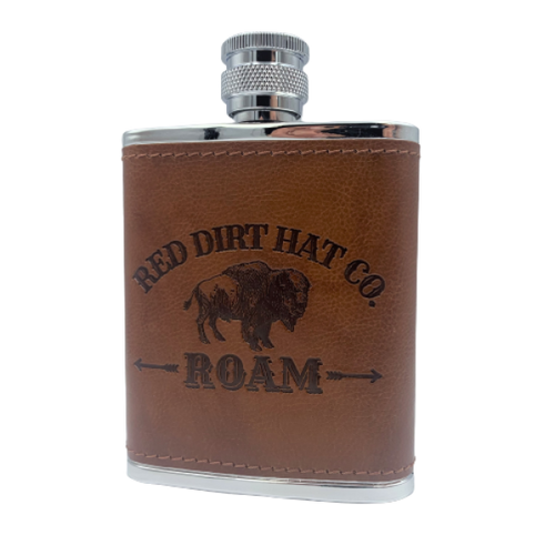 A bottle of Red Dirt Hat Company Roam cologne, with a brown leather-like wrap around the lower half and a metal cap. The bottle features the logo and the word 'ROAM' engraved on the front. It looks similar to a flask.