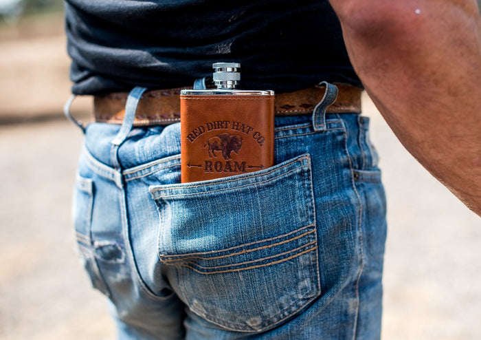 A bottle of Red Dirt Hat Company Roam cologne, with a brown leather-like wrap around the lower half and a metal cap. The bottle features the logo and the word 'ROAM' engraved on the front. It looks similar to a flask.