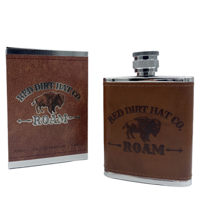A bottle of Red Dirt Hat Company Roam cologne, with a brown leather-like wrap around the lower half and a metal cap. The bottle features the logo and the word 'ROAM' engraved on the front. It looks similar to a flask.