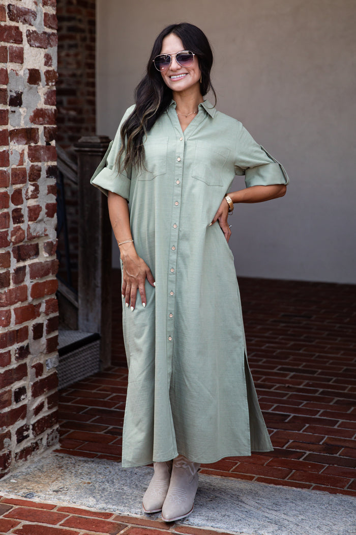 A woman wearing a sage green short-sleeve button-up maxi shirt dress, styled with beige ankle boots, sunglasses, and a gold watch, standing in front of a brick and stucco wall.