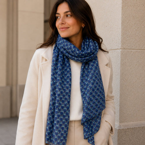 Woman wearing a blue patterned scarf and beige coat against a beige wall.