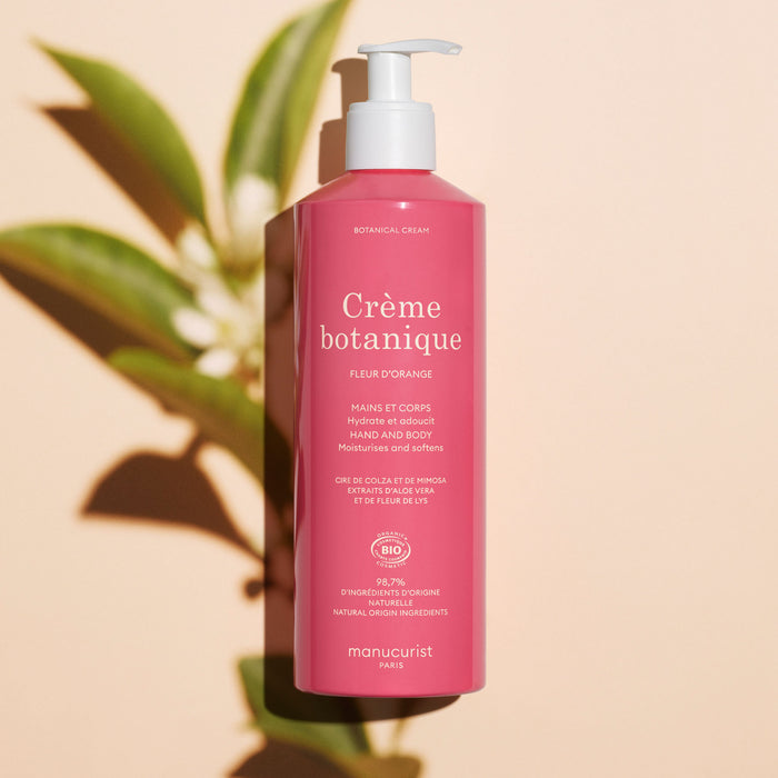 A pink bottle of Manucurist Soothing Orange Blossom Hand  Body Cream – Cosmos Organic with a pump dispenser stands on a light surface, casting a green leafy shadow. The bilingual label highlights its orange blossom fragrance.