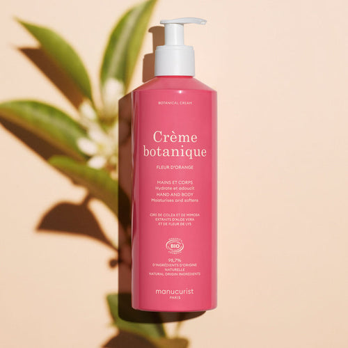 A pink bottle of Manucurist Soothing Orange Blossom Hand  Body Cream – Cosmos Organic with a pump dispenser stands on a light surface, casting a green leafy shadow. The bilingual label highlights its orange blossom fragrance.