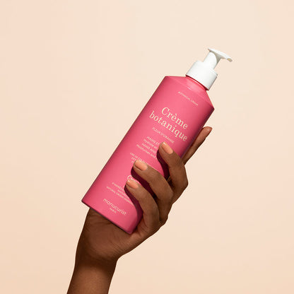 A hand holds a pink bottle of Manucurist Soothing Orange Blossom Hand  Body Cream - Cosmos Organic with a white pump dispenser against a plain beige background.