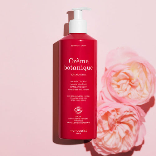 A pink bottle of Manucurist Soothing Rose Hand  Body Cream - Cosmos Organic stands upright on a light pink background beside two pink roses. The white pump and text highlight its promise to deeply hydrate your skin.