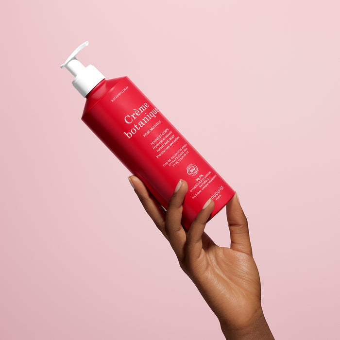A hand with dark skin holds up a bright red Manucurist Soothing Rose Hand  Body Cream - Cosmos Organic with a white pump, against a pale pink background. The label features white text, and the product is designed to deeply hydrate skin.