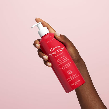 A hand holds a red bottle of Manucurist Soothing Rose Hand  Body Cream - Cosmos Organic with a white pump, against a light pink background. The label features white text highlighting its natural origin and deep hydration benefits.