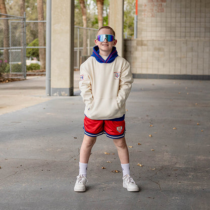 Smiling boy with hands in pockets wearing iridescent shield sunglasses a cream stadium hoodie and red mesh shorts