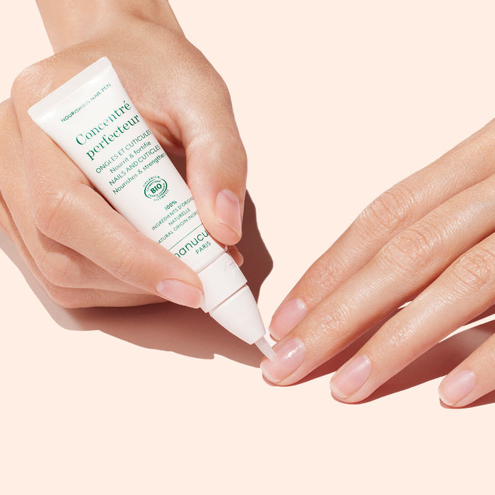 A person applies Strong Glow Trio nail treatment from Manucurist, using one hand to hold the tube while the other rests on a light pink surface for gentle nail care.
