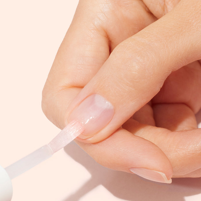 A close-up of a hand applying Manucurist Strong Glow Trio to a fingernail with a brush against a light beige background, emphasizing gentle nail care and treatment for damaged nails.