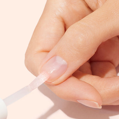 A close-up of a hand applying Manucurist Strong Glow Trio to a fingernail with a brush against a light beige background, emphasizing gentle nail care and treatment for damaged nails.