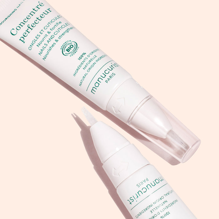 Two white manicure pens from the Stronger Nails Duo by Manucurist, shown on a peach background. One pen is closed and the other shows a brush tip. Both feature green text and are labeled “Concentré perfecteur.”.