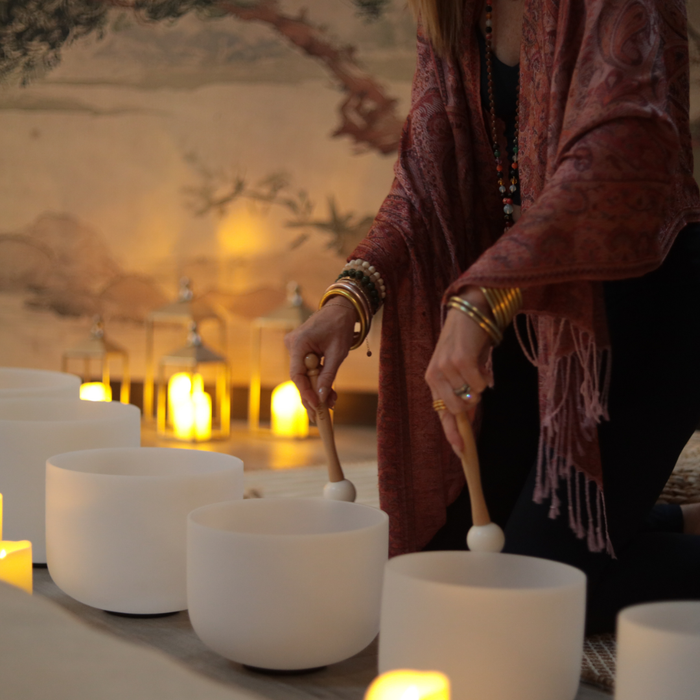 BuDhaGirl sound bath scene with a facilitator playing white singing bowls beside glowing candles in a warm tranquil setting