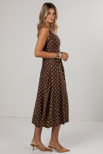 A woman modeling a sleeveless brown midi dress with a white polka dot pattern, paired with beige slingback heels, against a plain light gray studio background.