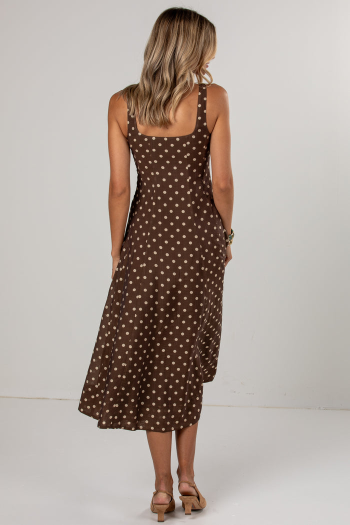 A woman viewed from behind wearing a brown midi dress with a white polka dot pattern, paired with tan heeled sandals, set against a plain light gray studio background.