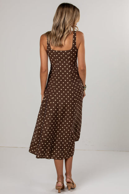 A woman viewed from behind wearing a brown midi dress with a white polka dot pattern, paired with tan heeled sandals, set against a plain light gray studio background.