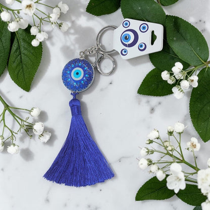 Blue evil eye keychain with tassel on a marble surface with white flowers and green leaves.