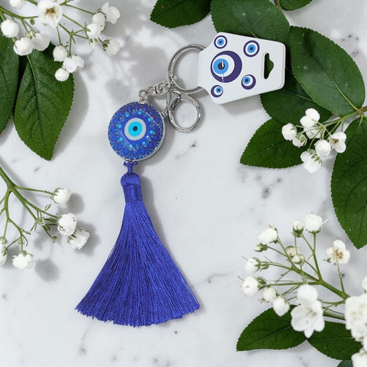 Blue evil eye keychain with tassel on a marble surface with white flowers and green leaves.