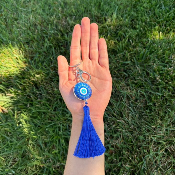 Hand holding a blue evil eye keychain with tassel against a grass background