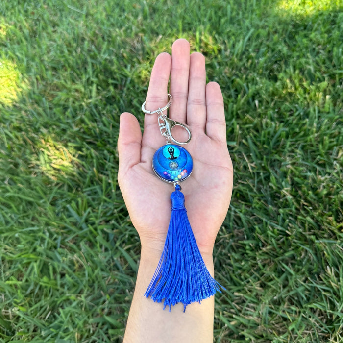 Hand holding a blue keychain with tassel against a grass background
