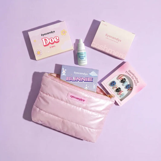 EyeCandys' The Complete Travel Pack, featuring a pink quilted pouch with the EyeCandys logo, displayed on a light purple surface alongside contact lens boxes, a bottle of contact lens solution, cat-themed lens cases, and a press-on nail set.