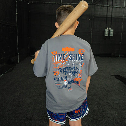 Child wearing a gray t-shirt with colorful graphics and text, holding a baseball bat in an indoor batting cage.