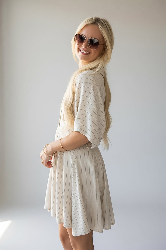 A blonde woman wearing a cream-colored pleated short-sleeve dress and sunglasses, posing in a studio setting against a light gray background.