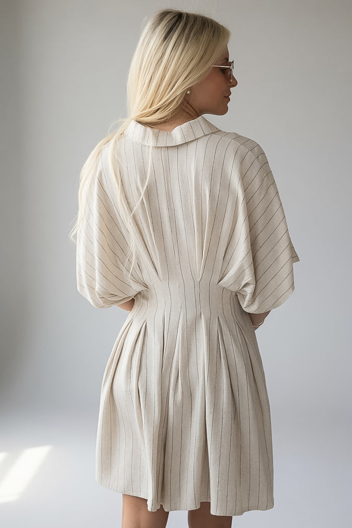 A woman with blonde hair is shown from the back wearing a pleated, short dress with wide three-quarter sleeves. The dress is a light cream or off-white color with thin vertical stripes, set against a plain light gray studio background.
