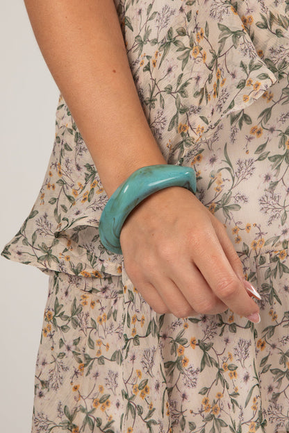 A close-up of a person's hand wearing a thick, glossy turquoise bangle, paired with a beige floral dress.