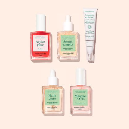 Five skincare products from Manucurist, including serums, oils, and a cream from the Ultimate Routine - Sensitive Nails collection, are arranged on a light pink background. Among them is a nourishing cuticle serum formulated to strengthen nails and enhance your nail care routine. Each product features a pastel-colored label highlighting its name and specific purpose.