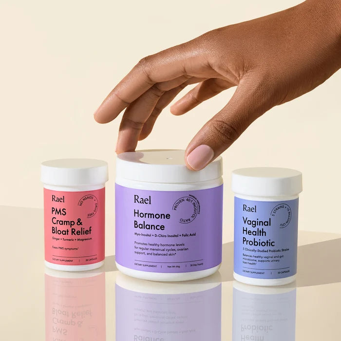 Hand about to grab one of three supplement jars labeled PMS Cramp & Bloat Relief, Hormone Balance, and Vaginal Health Probiotic by Rael.