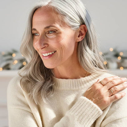 Smiling woman with long silver hair wearing clear under-eye gel patches and a cream sweater, with holiday lights behind her