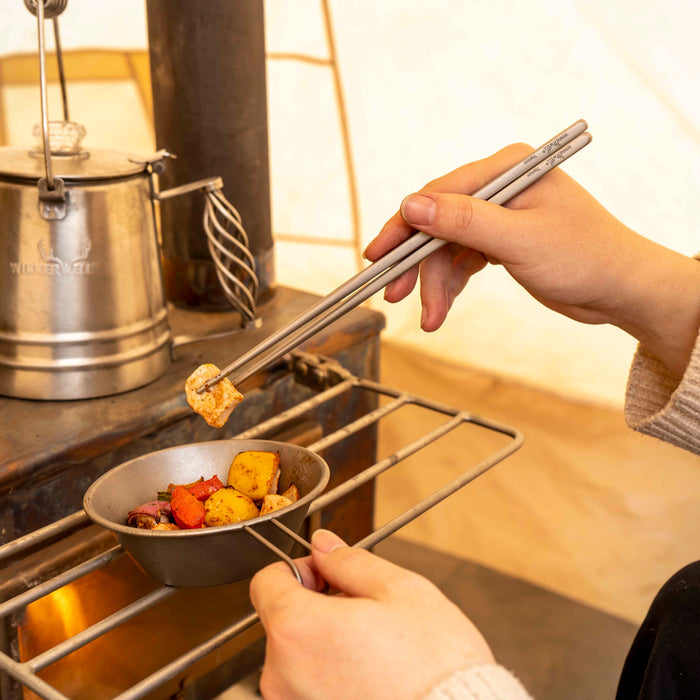 Hands using Winnerwell® square titanium chopsticks to cook food on a grill, demonstrating their practical application in outdoor cooking.