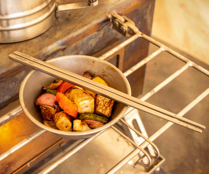 Winnerwell® square titanium chopsticks being used to cook a mix of ingredients on a grill, showcasing their use in outdoor culinary activities.