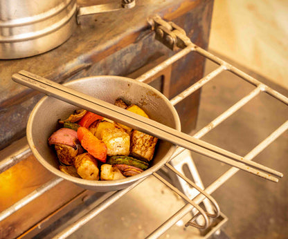 Winnerwell® square titanium chopsticks being used to cook a mix of ingredients on a grill, showcasing their use in outdoor culinary activities.
