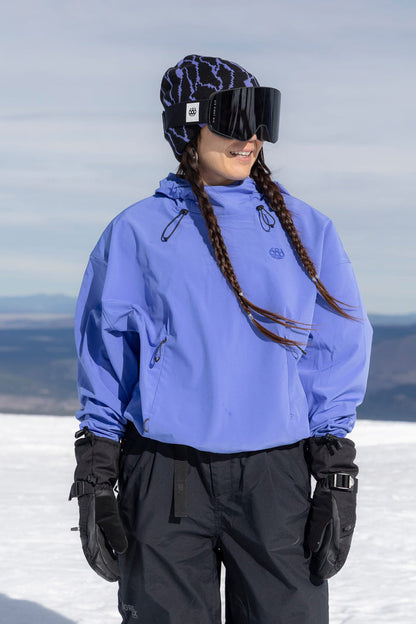 A woman wearing the 686 Women’s Waterproof Team Crop Hoody in blue, paired with black snow pants, gloves, large ski goggles, and a patterned beanie stands on the snow with braided hair, looking to the side. Mountains and a cloudy sky fill the background.