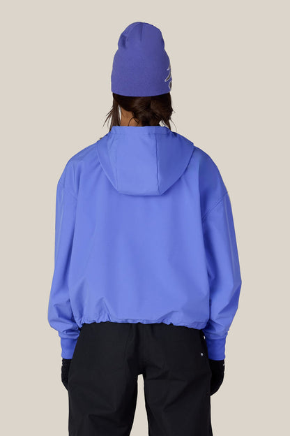 A person facing away from the camera is wearing the 686 Women’s Waterproof Team Crop Hoody in blue, paired with a matching blue beanie and black pants, set against a plain beige background.