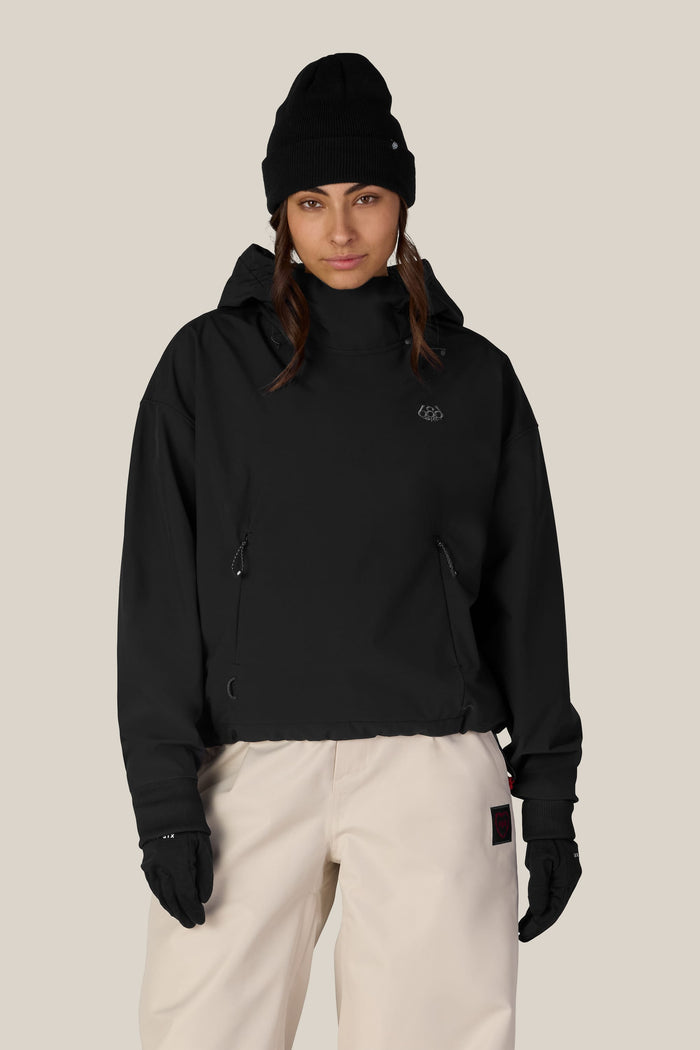A person wearing a black beanie, black gloves, white pants, and the 686 Women’s Waterproof Team Crop Hoody stands facing forward against a plain beige background.