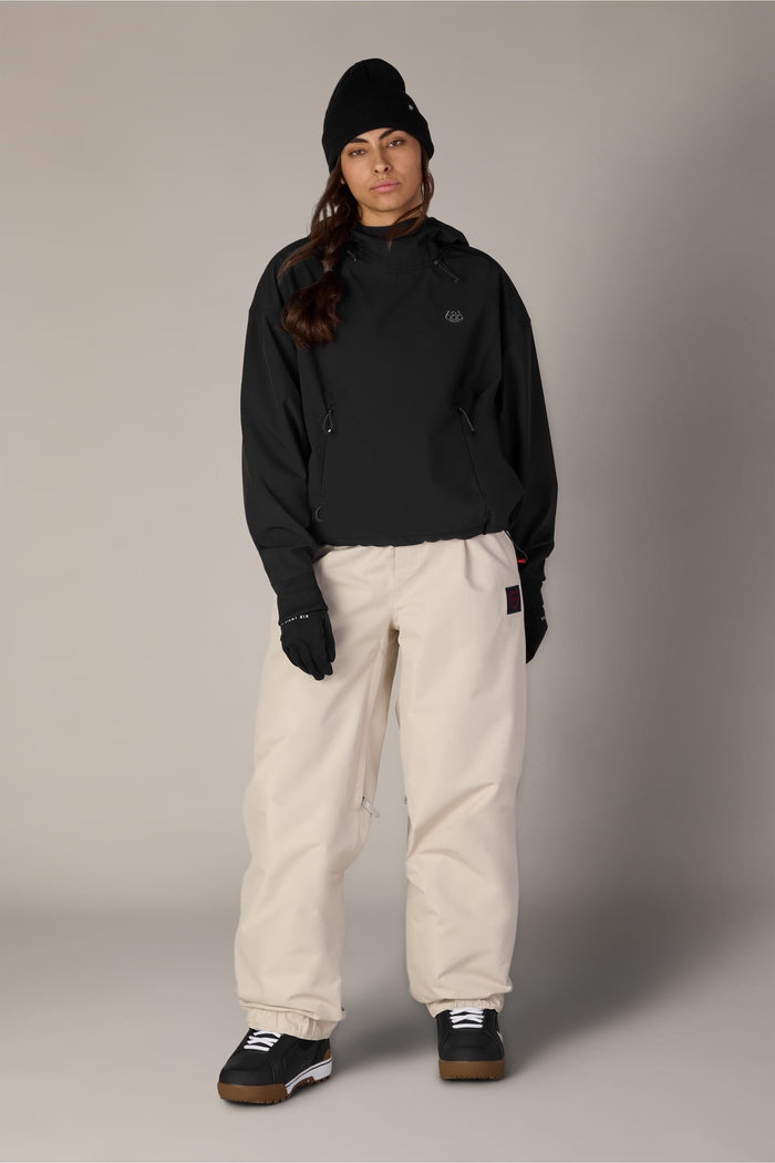 A person stands facing forward against a plain neutral background, wearing a black beanie, a black 686 Women’s Waterproof Team Crop Hoody layered under a black jacket, black gloves, beige baggy pants, and black sneakers with white soles.